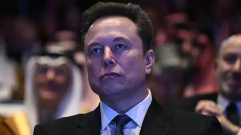 AFP via Getty Images Elon Musk, wearing a black suit, white shirt and dotted tie, looks away from the camera as he watches President Donald Trump speak at an event in Washington DC in November