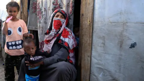 A woman wearing a niqab and holding a young child sits in a doorway as an older child stands next to them