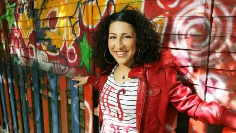 BBC A young woman with curly brown hair, wearing a red leather jacket and white-striped top, smiling at the camera and standing in front of a brightly graffitied wall
