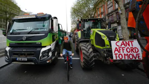 Brian Lawless/PA Wire A cyclist wearing an orange helmet is between two tractors. A sign says, can't afford to move. They're on a street. 