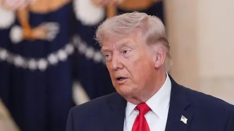 Getty Images US President Donald Trump speaks while wearing a dark suit, white shirt and red tie. He stands indoors front of flag with a the US presidential seal on it.