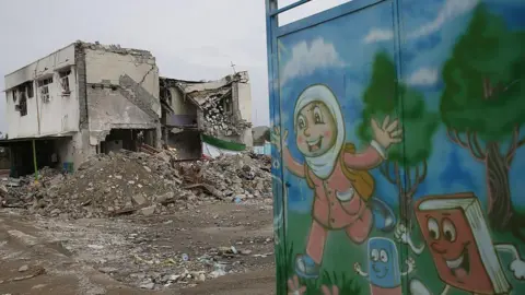 Anadolu via Getty Images A bombed-out school surrounded by rubble, with children's illustrations painted on an adjacent gate. 