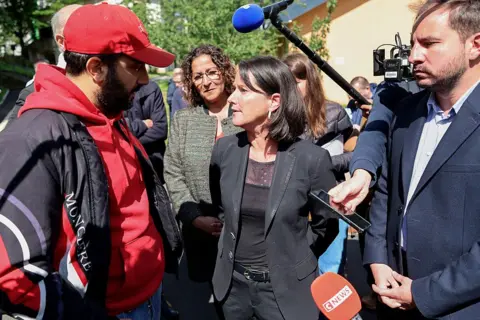 Getty Images Local mayor Johanna Rolland (C), wearing a black jacket, top and trousers, speaks to a man wearing a red cap and hoodie and a dark jacket. A microphone is held above her head and several other journalists are seen recording her remarks with phones and microphones.