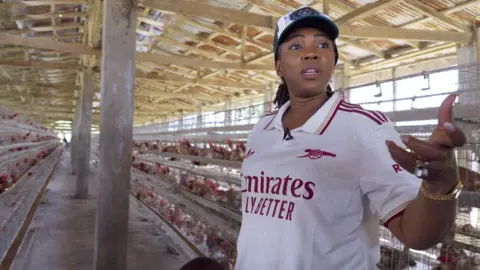 Fatima Bio inside a large chicken house lined with rows of cages. She wears a white Arsenal football shirt, a dark baseball cap, pearl earrings, and gold jewellery.
