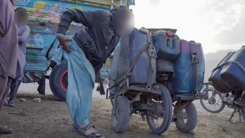 Irfan is pictured next to his bike, upon which cannisters of diesel have been strapped. He's wearing a dark blue jacket and light blue trousers. There's a van and a couple of fuel smugglers behind him