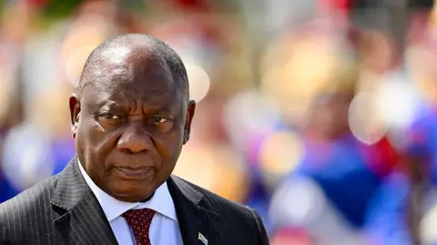 Getty Images A shot of President Cyril Ramaphosa, wearing a pin striped black suit with a white shirt and red patterned tie, from the shoulders up. He is looking off to the left and the background is blurred.