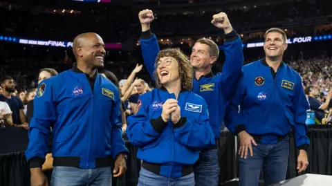 NASA The photo shows four astronauts from NASA’s Artemis II Moon mission standing in a busy indoor sports arena, wearing bright blue flight jackets with mission patches and logos. They are in the foreground, smiling and cheering, with one woman in the centre clasping her hands and two of the men raising their fists in the air in celebration. The crowd behind them is slightly out of focus but fills the stands, suggesting a major sporting event. The lighting is bright and even, and the mood is joyful, energetic and triumphant.