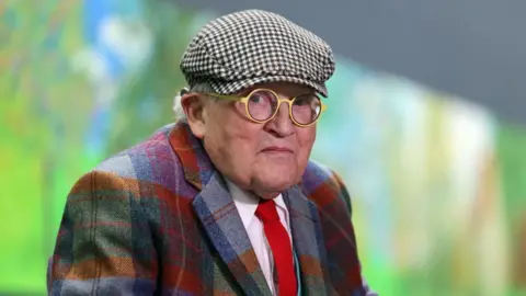 Getty Images British painter David Hockney poses at the Orangerie museum in Paris, on October 7, 2021, in front of his painting