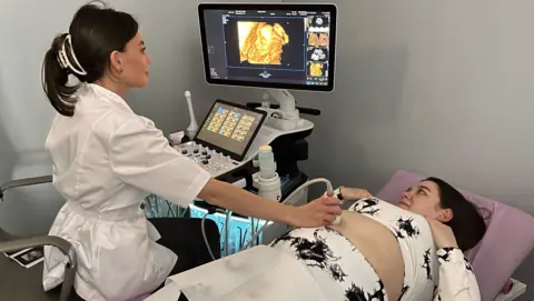 A pregnant woman is lying on a bed in a clinic having a scan. A woman in a white coat, with long, dark hair tied up, is carrying out the procedure. They are both looking at the screen, on which a foetus can be seen.