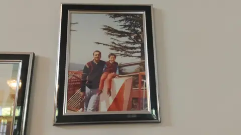 Kamal Saini/BBC Punjabi A picture frame of a man standing with his son in a hill station in India. The man is wearing a black sweater with red patches near the shoulder and has short hair and a mustache. His son - sitting right next to him - is wearing a maroon sweater vest with orange pants. 
