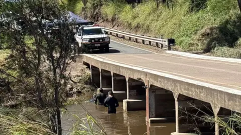 ABC Sunshine Coast: Jessica Ross Two men in wet suits wade in a brown river running underneath a concrete road bridge with a rescue vehicle and tent on the road