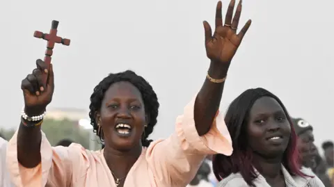 AFP via Getty Images A woman, in a pink top is smiling and holding both hands in the air. Her left hand is open, in a wave, and her right hand is holding a small wooden crucifix