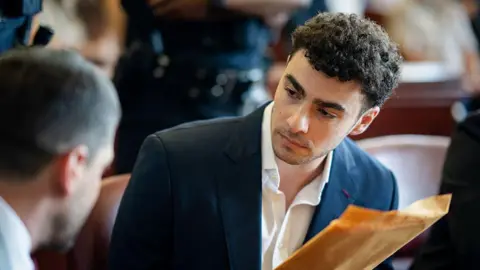 Getty Images Luigi Mangione looks toward his lawyer while wearing a navy jacket and white dress shirt. He is holding a yellow envelope in his hand.