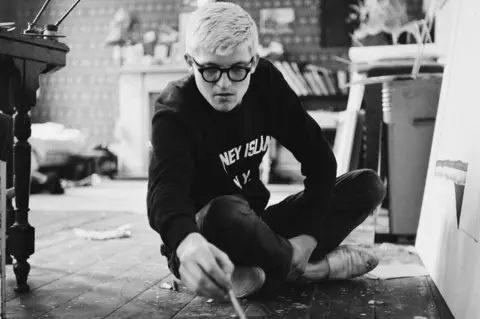 Getty Images David Hockney is seen in a black and white photo as a young man. He is intent on painting a canvas on the floor.