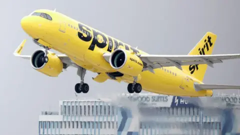 Getty Images A bright yellow aeroplane with Spirit's logos on the side and tail fin takes off from an airport with a hotel in the background.