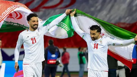 AFP via Getty Images Iran's players Omid Norafkan and Saman Ghoddos celebrate after a qualifying match. They are smiling and dressed in white football kits. One of them is waving an Iranian flag above his head.