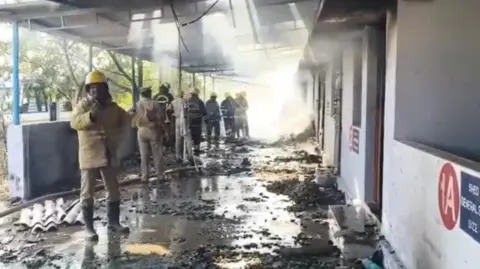 BBC Tamil A first responder in a khaki uniform and yellow helmet stands inside a lobby where a blast has occurred. Other officials gather in the background and debris lies scattered across the floor.