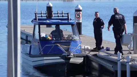 Reuters Two police officers stand on a jetty. One holds a large white box. Next to the jetty is parked a blue and white police boat, in which a third officer is sitting. All is surrounded by still and clear blue water