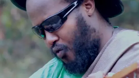 Eliel Sando via YouTube Close up of Karasira's face. He has a beard, is wearing sunglasses and is holding a musical instrument, just slightly visible in the bottom left corner. the rim of a hat and also be seen on either side of his head. His lips are pursed as he is singing