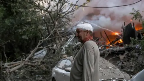 EPA A man wearing a grey top stands with his head bandaged as he surveys the aftermath of an attack. Beside him is a silver car, whose roof is covered with debris, trees that have fallen down in what appears to be a forested area and where a fire is still burning bright