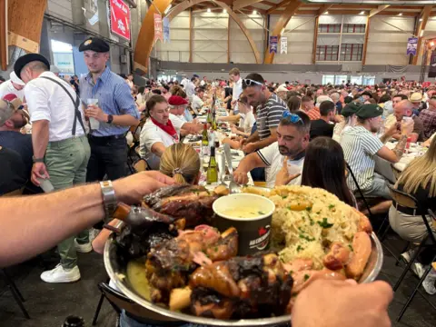 In the foreground of the image is a large plate of meat and potatoes. It is being held on a silver platter by a man with a metal wrist watch. In the background hundreds of people, sit at long tables there a bottles of wine down the centre of the tables