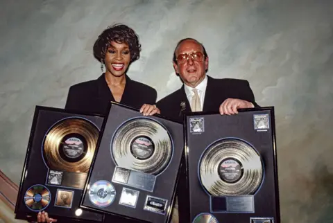 Getty Images Whitney Houston and Clive Davis hold up a collection of gold and platinum records, celebrating the success of her album I'm Your Baby Tonight