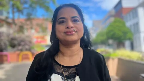 Biji Jose is standing in a street and looking into the camera. She's wearing a black jacket and her top is black and white with lace.