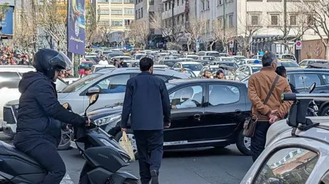 Getty Images Iranians try to clear a street amid heavy traffic in Tehran, Iran, on February 28, 2026. One man on a motor scooter and two others on foot stand around with rows of stationary cars in the background on a city street.