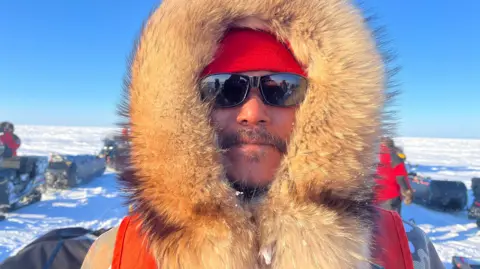 Eloise Alanna/BBC Barnie Aggark, an Inuk man, pictured wearing a hood with a thick fur trim and sunglasses. (for the photo captions). 