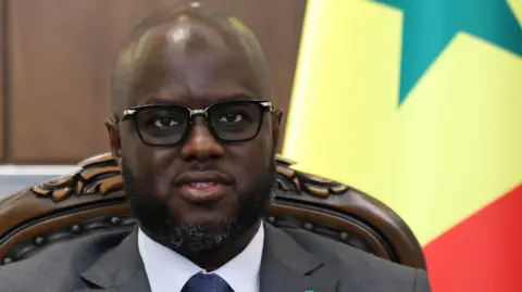 Anadolu via Getty Images El Malick Ndiaye wears a suit and tie and sits in front of the Senegalese national flag.
