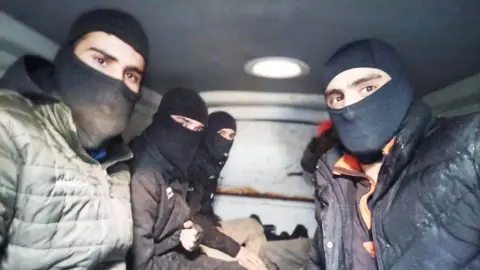 Supplied to BBC A group of men wearing black balaclavas sit close together in the back of a dimly lit van. Each person wears a heavy winter jacket and they're all looking intently into the lens of the camera.