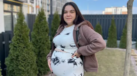BBC A pregnant woman stands in a garden with her hand on her belly. She has long, straight, dark hair and is wearing a matching top and trousers which are white with some patches of black. She also has a light brown hoodie on. 