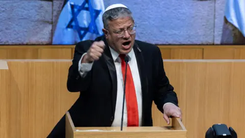 Reuters Itamar Ben-Gvir wearing a black suit, red tie and white kippah, talking on a podium in front of Israeli flags