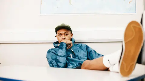 Beyond Equality/ Ben Hurst Ben Hurst sat at a table wearing an olive cap, blue hoodie, feet crossed and perched on a desk, looking directly at the camera posing with his hand over his mouth.