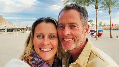 Free Lindsay and Craig Campaign A couple on a beach. It is sunny and the sand is white. Behind them are some trees.