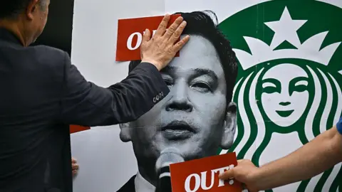AFP via Getty Images Activists put red stickers stating "out!" on an image of Shinsegae Chairman Chung Yong-jin during a rally calling for a boycott of the Starbucks coffee chain in Seoul last month.
