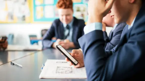 Getty Images Students in school looking at their phones. We can see two students from the side in a navy school uniform and one student further away facing the camera who is blurred