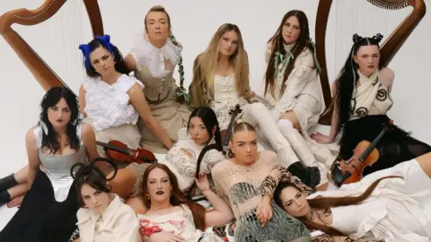 Cat Couture A group of eleven female musicians pose together in a studio setting, wearing elaborate and varied neutral toned outfits, arranged around two large harps with several holding traditional instruments such as fiddles.