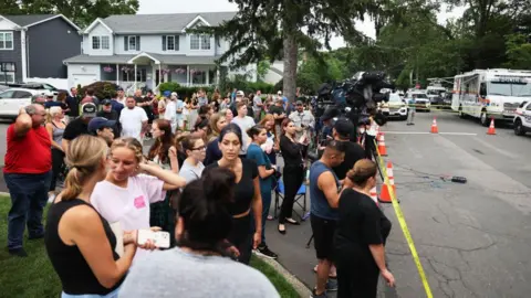 Getty Images A crowd assembles on a suburban street behind police tape and police vehicles