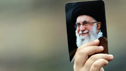 EPA/Shutterstock Just a hand can be seen, the rest of the person out of shot - holding up a picture of the late Iranian Supreme Leader Ayatollah Ali Khamenei. He is an elderly man with a white beard, pictured smiling. The portrait is about the size of a playing card.