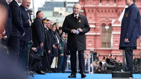 AFP via Getty Images Vladimir Putin walks on a stage wearing a suit with a red building in the background