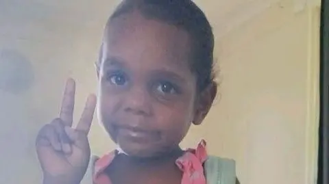 Northern Territory Police An girl wearing a pink dress and blue backpack poses for a picture, holding up the peace sign