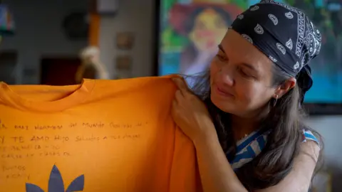 Adriana Briceño holds up an orange t-shirt with a message written on it in pen 