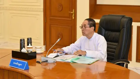 Office of the Commander-in-Chief of Defence Services Myint Swe in a white long-sleeved shirt sitting at a large table, with documents laid out in front of him