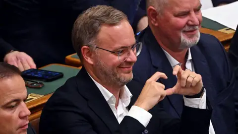 AFP via Getty Images Balázs Orbán makes a sign with his fingers as he grins in parliament