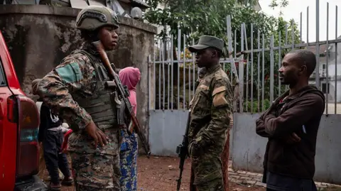 AFP via Getty Images M23 members, in military uniform and each with a guns, stand guard near a private residence in Goma