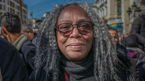 NurPhoto A person with grey, shoulder‑length hair in a crowded street during a march organised by the Tunisian General Labour Union (UGTT) in Tunis, surrounded by other participants and urban buildings in the background.