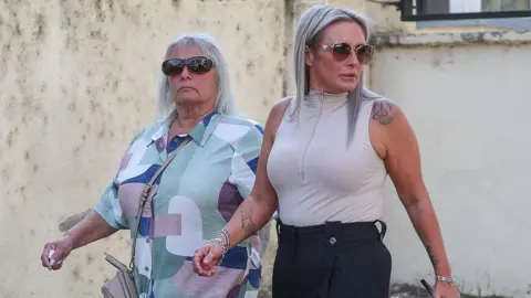 Reuters Bella Culley's mother, Lyanne Kennedy, and, on her right, Bella's grandmother, Christine Cook, walking outside the Tbilisi City Court building in Tbilisi in Georgia. Lyanne Kennedy has long, dyed platinum-grey-blonde hair with dark roots showing and is wearing silver necklaces, bracelets and rings, and has on sunglasses. She is wearing smart, black trousers and a pale, pinky-grey sleeveless top with a front zip, zipped up, and has tattoos on her left shoulder and right forearm. Christine Cook is wearing a blue, green, white and purple geometric design shirt and has shoulder length, wispy, white hair. She is wearing dark sunglasses and has a stern expression.