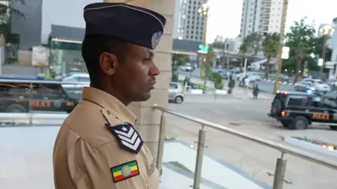 BBC A sideview of a policeman in beige uniform with a dark blue cap, an Ethiopian flag badge and rank insignia are on his shoulder. A main road can be seen behind him.