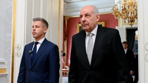 EPA Peter Magyar and Tamas Sulyok walking through a door. A chandelier hags in the room behind them, which also has a room with red-coloured wallpaper. 